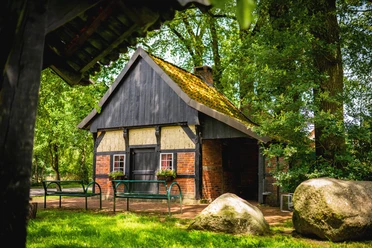 Backhaus-Heitel_Lünne©MedienAtelierEmsland.jpg Historisches Backhaus mit Fachwerkwänden, Ziegeldach und grüner Sitzbank unter hohen Bäumen.