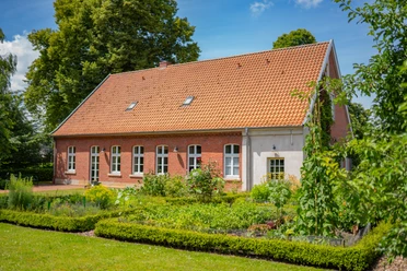 Haus-Schmeing_Lünne©MedienAtelierEmsland.jpg Backsteingebäude mit rotem Ziegeldach, umgeben von gepflegtem Garten und hohen Bäumen im Sommer.