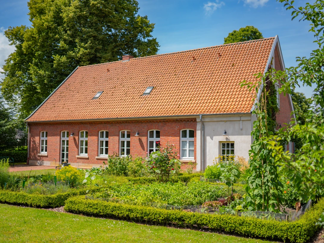 Haus-Schmeing_Lünne©MedienAtelierEmsland.jpg Backsteingebäude mit rotem Ziegeldach, umgeben von gepflegtem Garten und hohen Bäumen im Sommer.