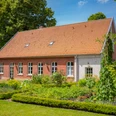 Haus-Schmeing_Lünne©MedienAtelierEmsland.jpg Backsteingebäude mit rotem Ziegeldach, umgeben von gepflegtem Garten und hohen Bäumen im Sommer.