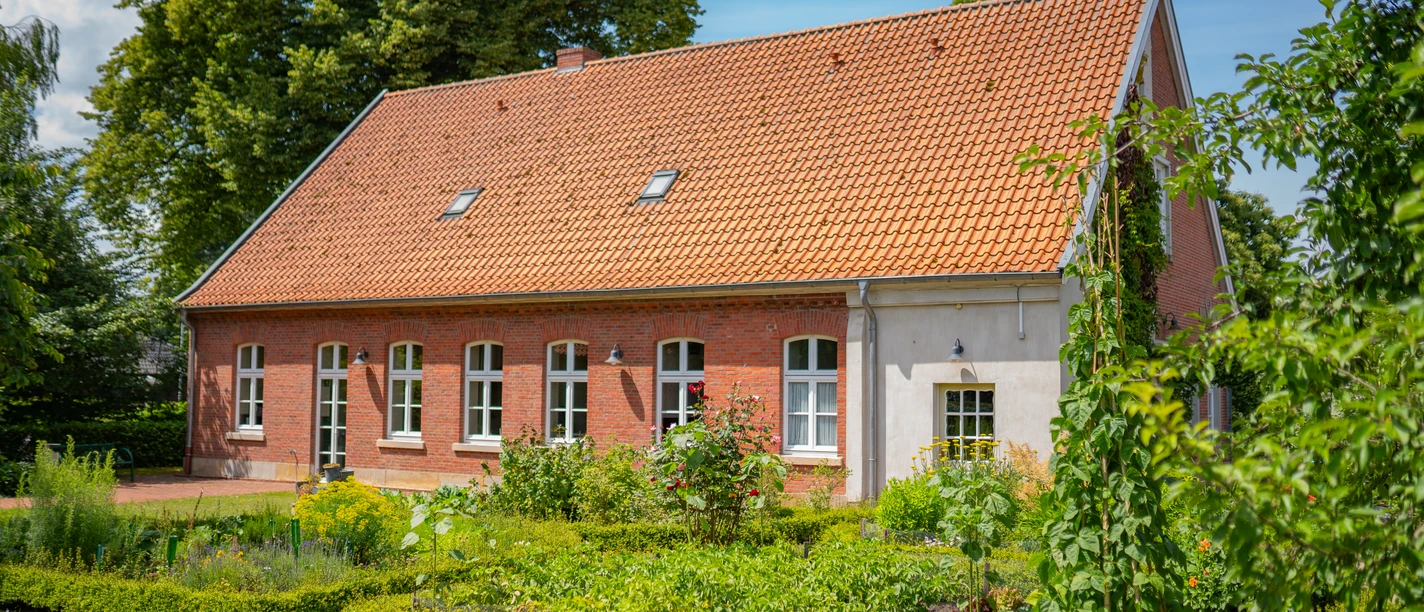 Haus-Schmeing_Lünne©MedienAtelierEmsland.jpg Backsteingebäude mit rotem Ziegeldach, umgeben von gepflegtem Garten und hohen Bäumen im Sommer.