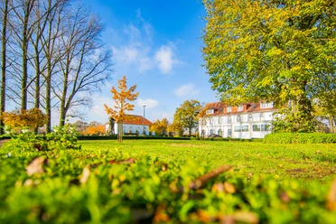 Ehem.-Handelsschule_Schapen©MedienAtelierEmsland.jpg Weißes historisches Gebäude mit rotem Dach hinter grüner Parkwiese und buntem Laub im Sonnenlicht.