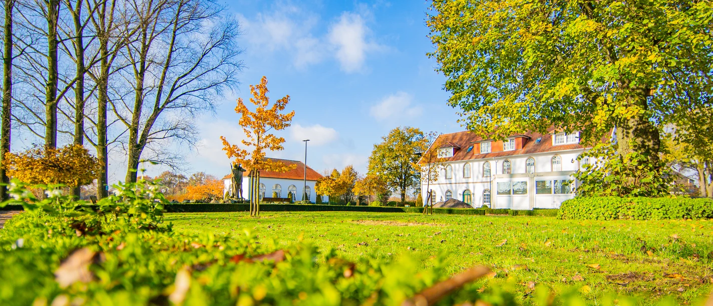 Ehem.-Handelsschule_Schapen©MedienAtelierEmsland.jpg Weißes historisches Gebäude mit rotem Dach hinter grüner Parkwiese und buntem Laub im Sonnenlicht.