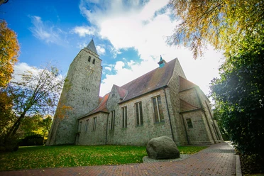 Kath.Kirche-St.Ludgerus_Schapen©MedienAtelierEmsland.jpg Historische Kirche aus hellem Naturstein mit Glockenturm und rotem Ziegeldach unter blauem Himmel