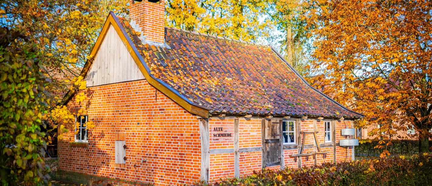 Alte-Schmiede-Laake_Varenrode©MedienAtelierEmsland.jpg Backsteinbau mit Ziegeldach und Schornstein, umgeben von herbstlichen Bäumen in warmem Licht