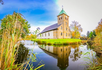 Pfarrkirche-St.Vitus-Venhaus©MedienAtelierEmsland.jpg Steinkirche mit grünem Turmdach am Wassergraben, umgeben von Bäumen unter blauem Himmel.