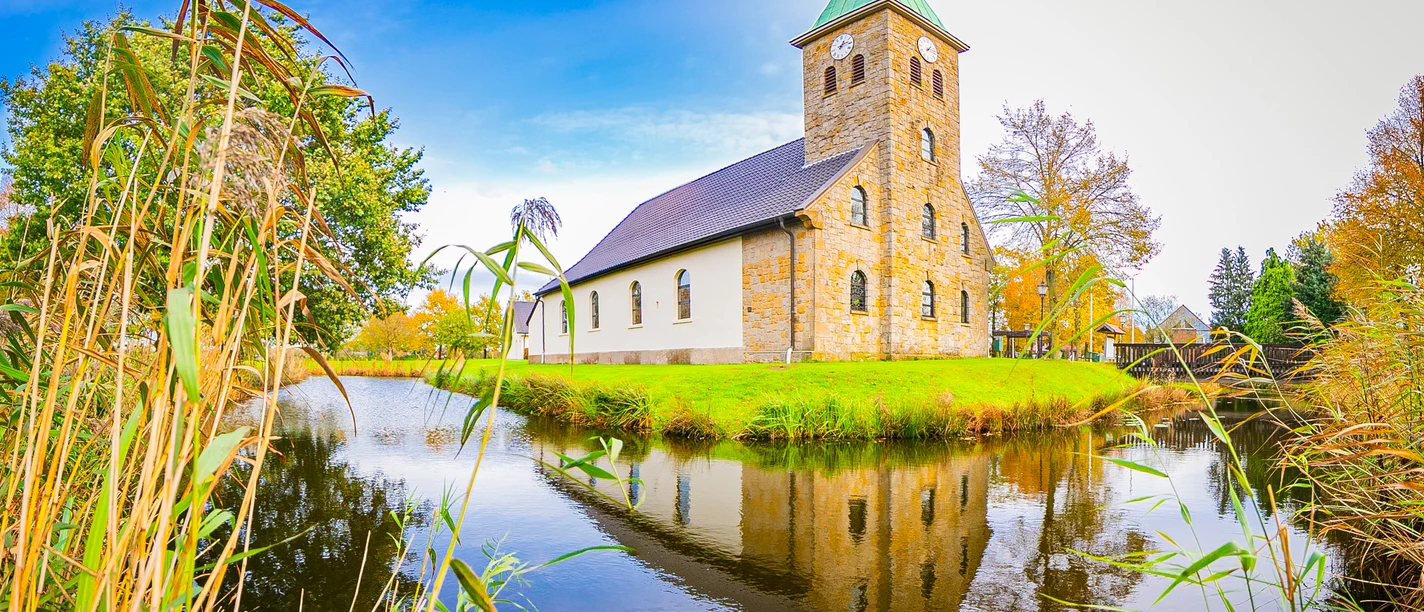 Pfarrkirche-St.Vitus-Venhaus©MedienAtelierEmsland.jpg Steinkirche mit grünem Turmdach am Wassergraben, umgeben von Bäumen unter blauem Himmel.