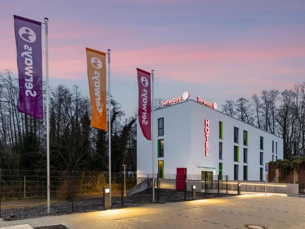Serways Hotel Hösel in Ratingen Mehrfarbige Fahnen wehen vor einem modernen rechteckigen Serways Hotel mit beleuchtetem Logo.
