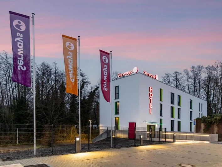 Serways Hotel Hösel in Ratingen Mehrfarbige Fahnen wehen vor einem modernen rechteckigen Serways Hotel mit beleuchtetem Logo.
