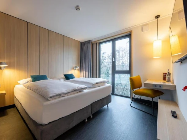 Hotelzimmer im Serways Hotel Hösel in Ratingen Modernes Hotelzimmer mit zwei Einzelbetten, helle Holzwände, Schreibtisch, Stuhl und Blick ins Freie.