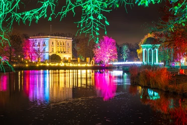 Christmas Garden Markkleeberg Das Bild zeigt eine farbenfrohe, nächtliche Szenerie mit festlich beleuchteten Bäumen und einem historischen Gebäude, das sich in einem ruhigen Gewässer spiegelt. Die intensiven Lichter und Farben verleihen der Umgebung eine magische Atmosphäre.