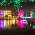 Christmas Garden Markkleeberg Das Bild zeigt eine farbenfrohe, nächtliche Szenerie mit festlich beleuchteten Bäumen und einem historischen Gebäude, das sich in einem ruhigen Gewässer spiegelt. Die intensiven Lichter und Farben verleihen der Umgebung eine magische Atmosphäre.