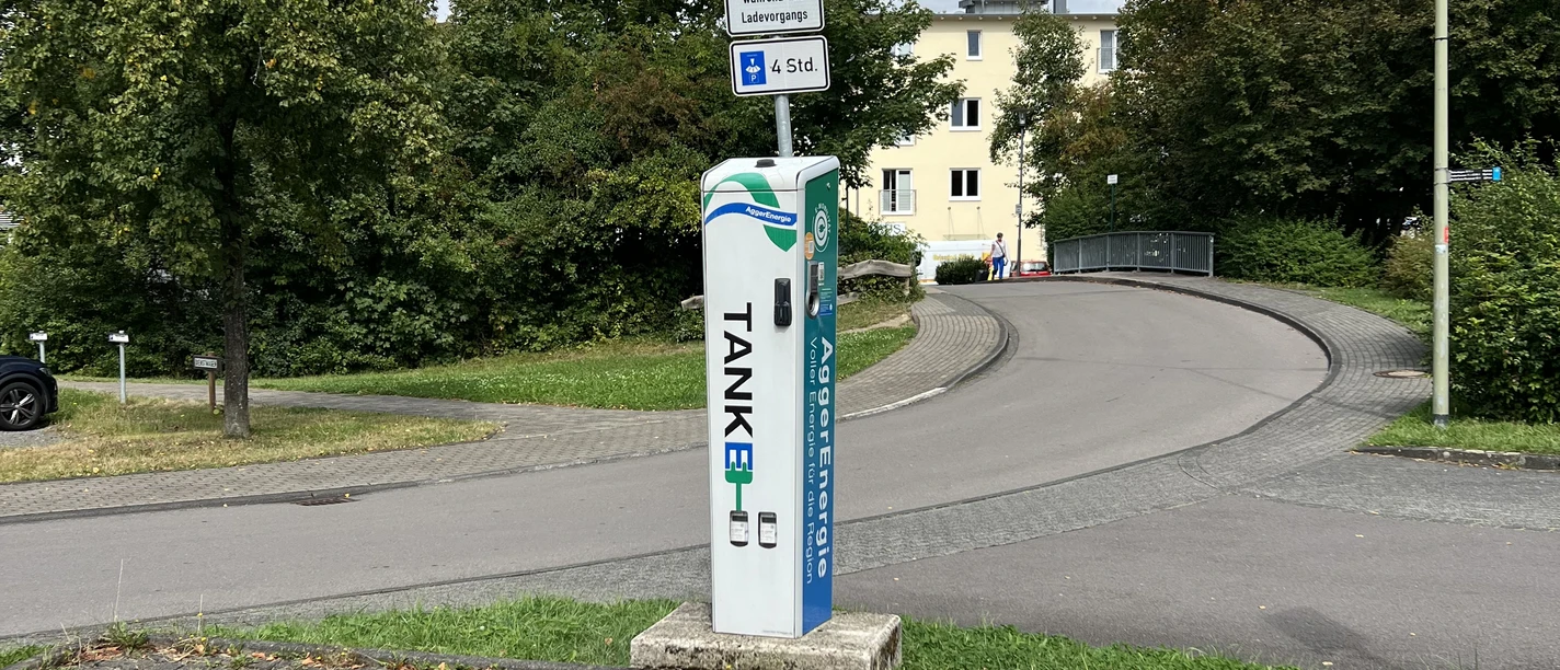 Ladesäule Parkplatz mit Ladestation für Elektrofahrzeuge, umgeben von Bäumen und einem Mehrfamilienhaus.