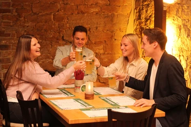 Barres Brauwelt Vier junge Erwachsene stoßen fröhlich mit Biergläsern in einer rustikalen Brauerei an.