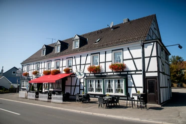 Restaurant Zum Musikalischen Wirt Fachwerkhausgasthof mit roten Blumenkästen am sonnigen Straßencafé, blauer Himmel im Hintergrund.