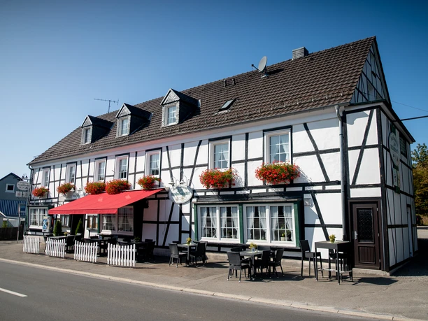 Restaurant Zum Musikalischen Wirt Fachwerkhausgasthof mit roten Blumenkästen am sonnigen Straßencafé, blauer Himmel im Hintergrund.