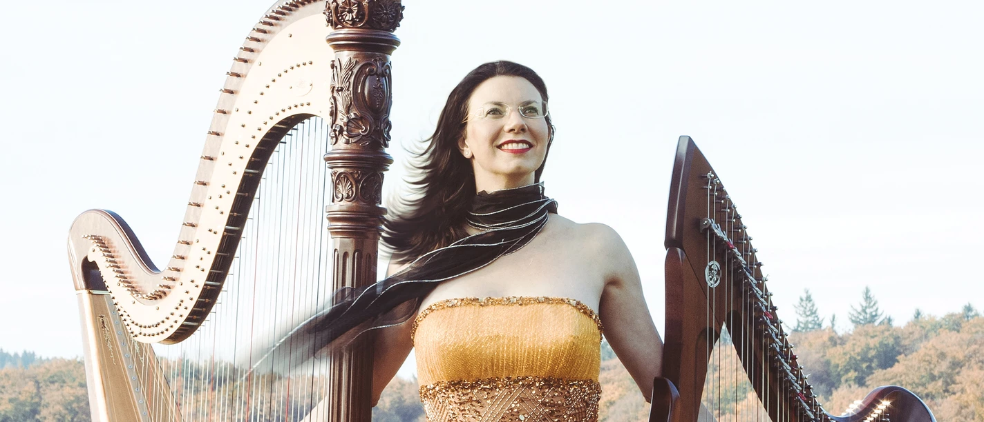 Bad Driburg_Musikgesellschaft_zwei harfen.jpg Eine Frau in einem eleganten, goldenen Kleid steht zwischen zwei Harfen auf einer Wiese vor herbstlichem Wald.