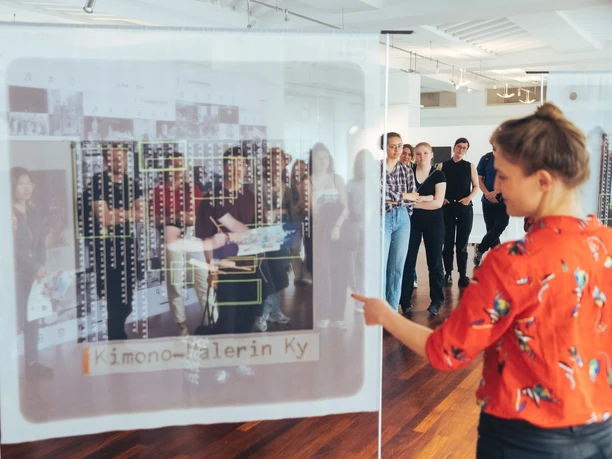 Group viewing translucent photo panels in a gallery as a woman gestures toward an artwork
