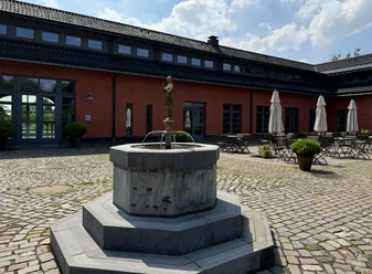 Malteser Kommende - Außenansicht Ein steinerner Brunnen in einem gepflasterten Hof vor einem roten Gebäude mit leeren Café-Tischen.