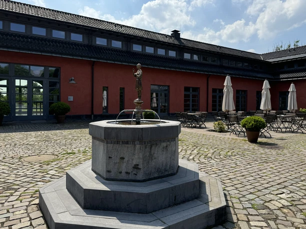 Malteser Kommende - Außenansicht Ein steinerner Brunnen in einem gepflasterten Hof vor einem roten Gebäude mit leeren Café-Tischen.