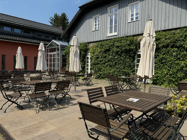 Malteser Kommende - Ansicht Außenbereich Innenhof mit Holztischen, Stühlen und Sonnenschirmen, umgeben von Gebäuden mit Kletterpflanzen.