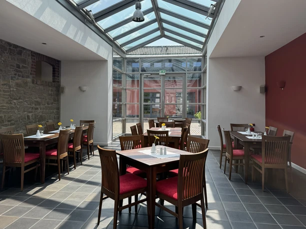 Malteser Kommende - Ansicht Frühstückssaal Gemütlicher Wintergarten eines Restaurants mit Holztischen, roten Stühlen und großem Glasdach.