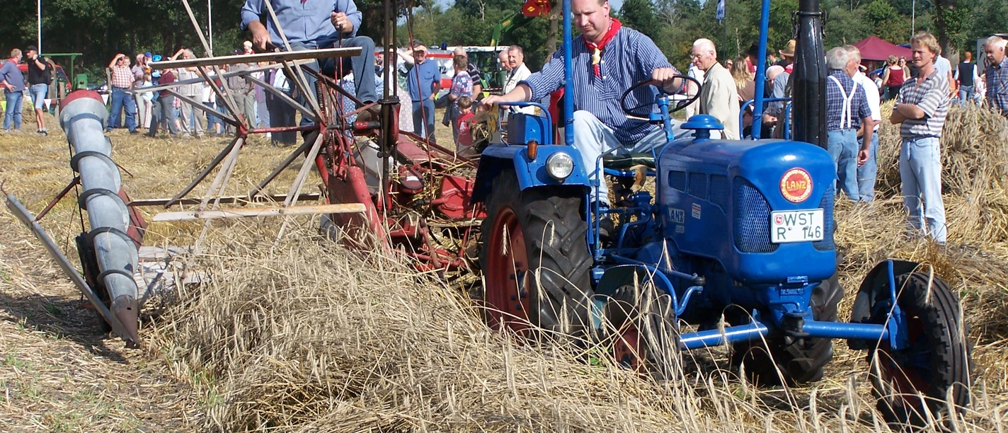 aper-berg-008 Ein Mann fährt einen blauen Lanz-Traktor durch ein abgeerntetes Getreidefeld. Dahinter sitzt ein weiterer Mann auf einem roten Mähwerk. Viele Menschen stehen am Feldrand und beobachten das Geschehen