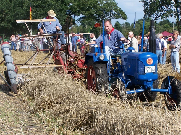 aper-berg-008 Ein Mann fährt einen blauen Lanz-Traktor durch ein abgeerntetes Getreidefeld. Dahinter sitzt ein weiterer Mann auf einem roten Mähwerk. Viele Menschen stehen am Feldrand und beobachten das Geschehen