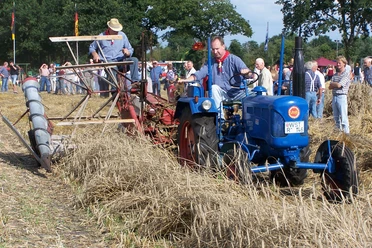 aperberg Ein Mann fährt einen blauen Lanz-Traktor durch ein abgeerntetes Getreidefeld. Dahinter sitzt ein weiterer Mann auf einem roten Mähwerk. Viele Menschen stehen am Feldrand und beobachten das Geschehen
