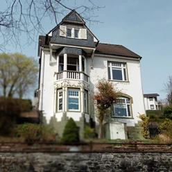 Fewo Villa Neviges in Velbert-Neviges Weiße, zweistöckige Villa mit dunklem Dach, großen Fenstern und dekorativen Balkonen umgeben von Grün.