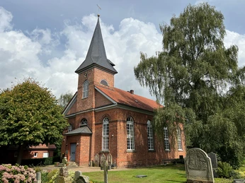 Backsteinkirche in Oiste mit spitzem Turm, umgeben von Bäumen und historischen Grabsteinen.