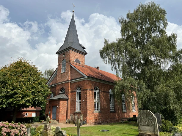 A brick church in Oiste with a pointed tower, surrounded by trees and historic gravestones.