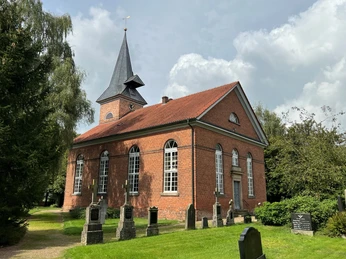 Backsteinkirche mit hohen Fenstern und spitzem Turm, umgeben von Rasen und alten Grabsteinen.