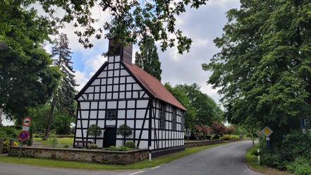 Fachwerk-Kapelle mit rotem Ziegeldach in grüner Umgebung; ruhige Straße führt links vorbei.