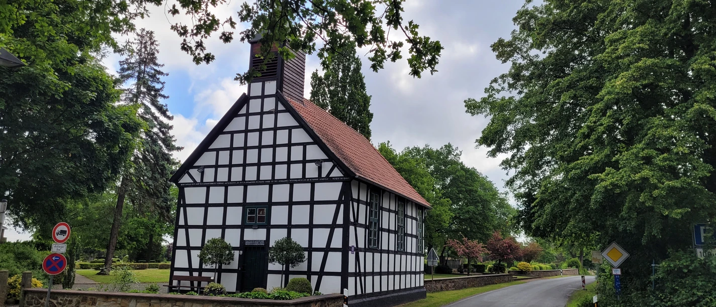 Alte Klus Espelkamp Frotheim Fachwerk-Kapelle mit rotem Ziegeldach in grüner Umgebung; ruhige Straße führt links vorbei.