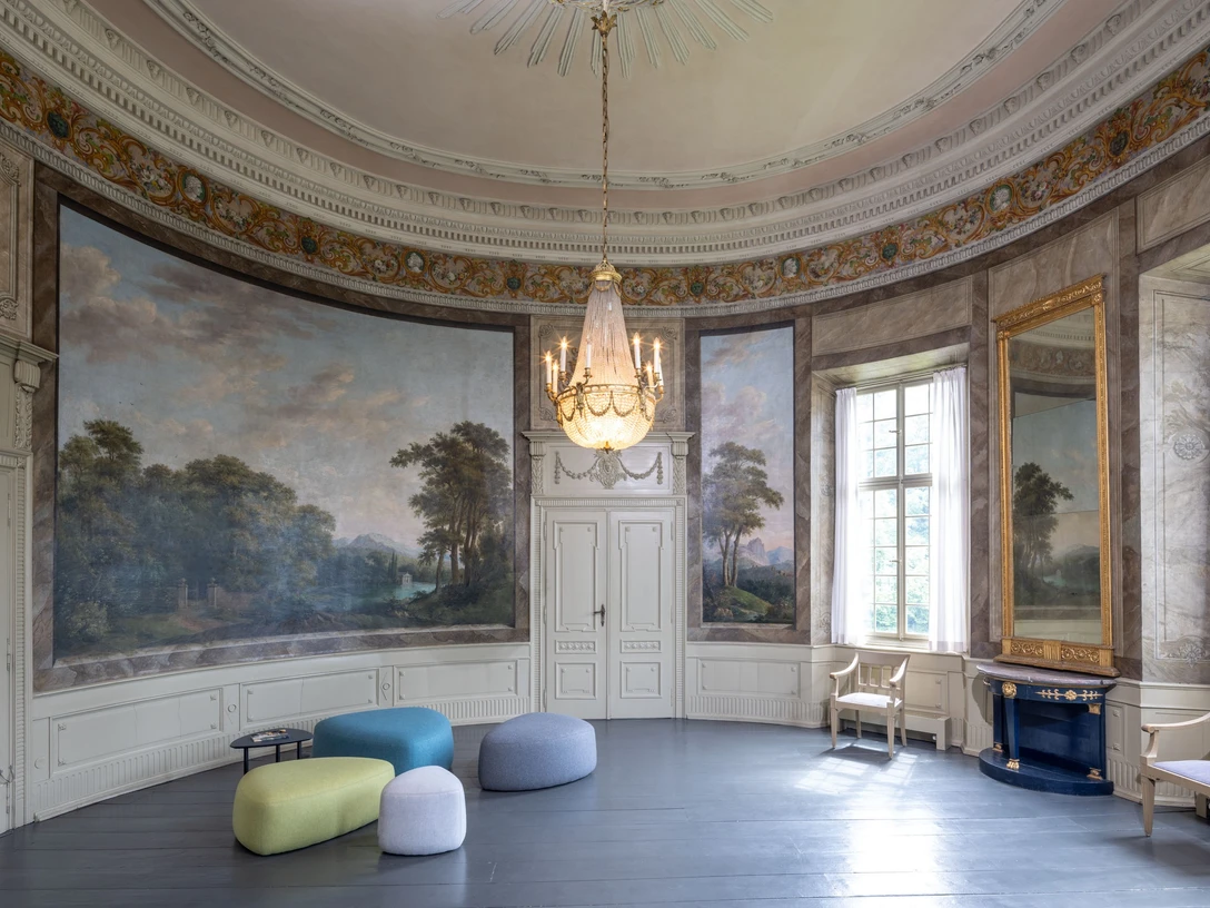 Gartensaal_Copyright_LVR-ZMB, Stefan Arendt 2024.jpg Eleganter, runder Saal mit historischen Wandmalereien, imposantem Kronleuchter und modernen Sitzhockern.