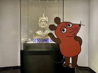 Ein hell erleuchteter Raum mit einer goldenen Büste von Karl dem Großen in einer gläsernen Vitrine. Daneben steht eine fröhliche Zeichentrickfigur, die Maus aus der Sendung "Die Sendung mit der Maus".