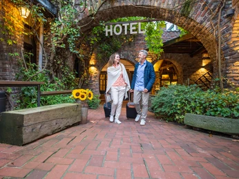 Mettmann Land Gut Höhne Ein Paar mit Gepäck geht auf einem Kopfsteinpflasterweg unter gewölbten, bewachsenen Torbögen.