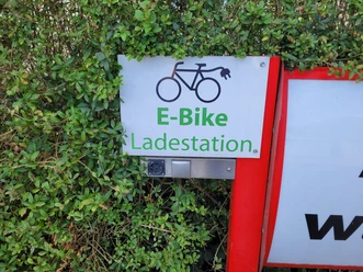 E-Bike Ladestation Dörpen ©Emsland Tourismus GmbH (1).jpg Schild einer E-Bike Ladestation an rotem Rahmen, teils von grünem Busch umgeben, mit Steckdose darunter.