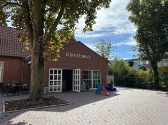 Klönstoawe Dorfladen in Brual ©Emsland Tourismus GmbH (1).JPEG Backsteingebäude mit geöffneten weißen Türen, Baum im Vordergrund und roter Kinderrutsche davor.