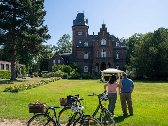 Monheim Marienburg Eine historische Backsteinvilla mit einem hohen Turm, umgeben von gepflegtem Rasen und Bäumen.