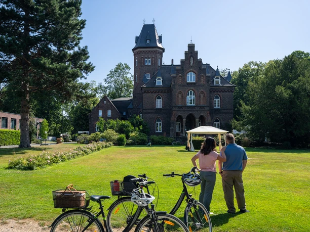 Monheim Marienburg Eine historische Backsteinvilla mit einem hohen Turm, umgeben von gepflegtem Rasen und Bäumen.