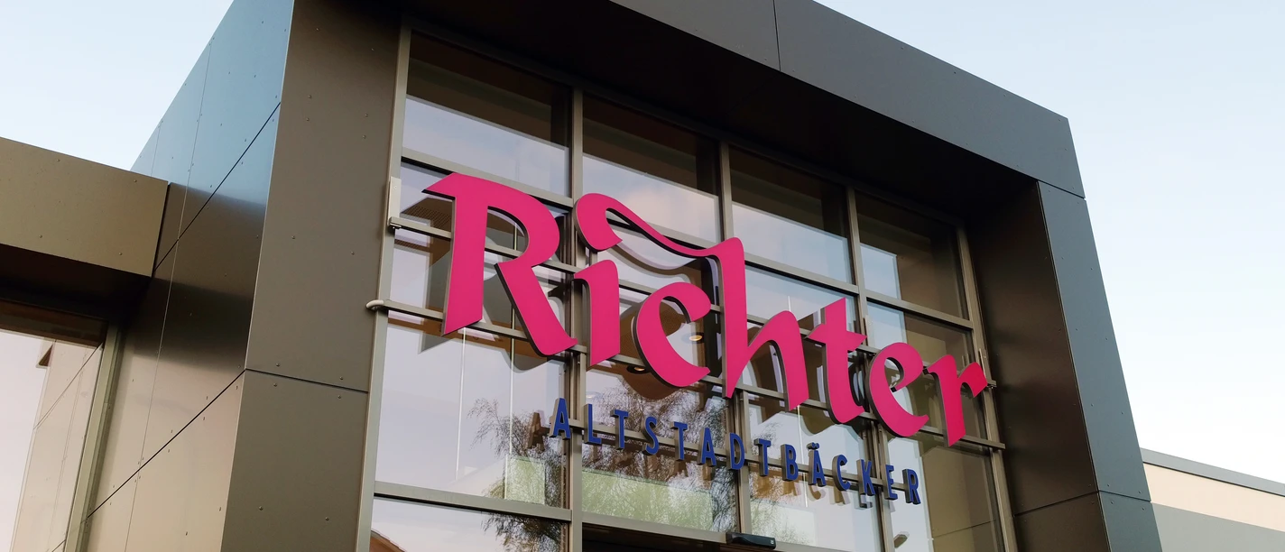 Close-up of an angular façade with large glass surfaces. Pink inscription: "Richter" underneath in blue: "Altstadtbäcker"