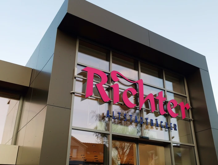 Eingang BACK.HAUS Nahaufnahme einer eckigen Fassade mit großen Glasflächen. Pinke Aufschrift: "Richter" darunter in blau: "Altstadtbäcker"