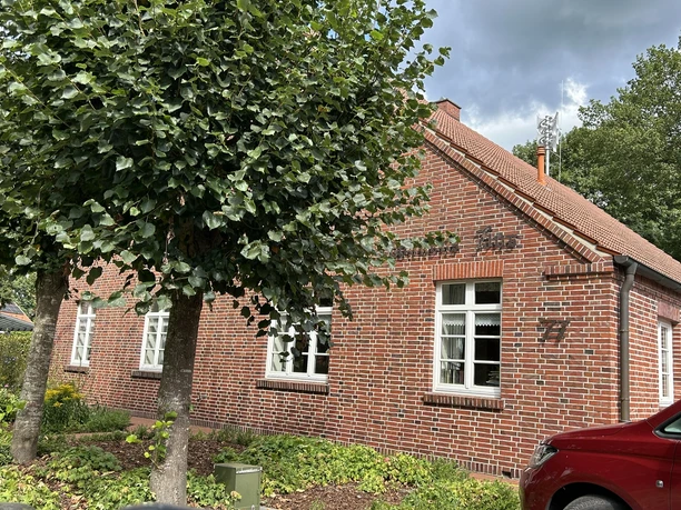 Heimathaus Hilkenbrook ©Emsland Tourismus GmbH (2).JPEG Backsteingebäude mit Satteldach, weißen Fenstern, Vorgarten und Bäumen unter bedecktem Himmel.