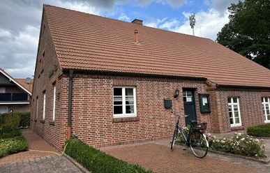 Heimathaus Hilkenbrook ©Emsland Tourismus GmbH (3).JPEG Backsteinhaus mit rotem Ziegeldach, grüner Tür, blühenden Rosen und abgestelltem Fahrrad davor.