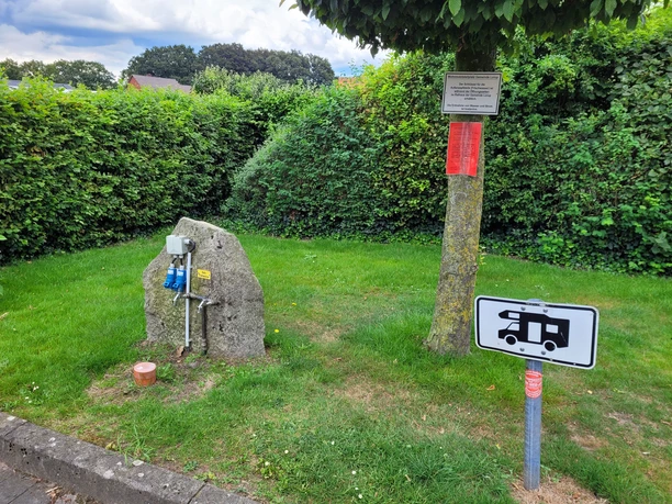 Stellplatz Rathaus Lorup ©Emsland Tourismus GmbH (2).jpg Grüne Stellfläche mit Steinblock, Stromanschlüssen und Wohnmobilschild vor dichter Hecke