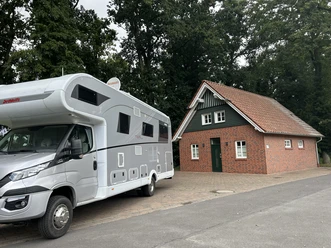 Großes Wohnmobil auf gepflastertem Stellplatz neben kleinem Backsteinhaus mit rotem Dach und Bäumen.