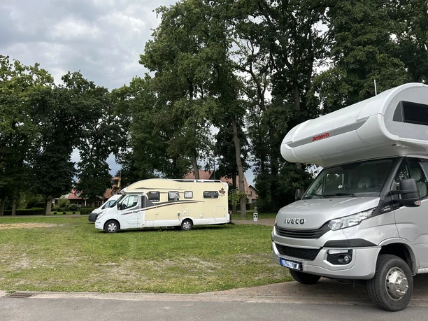 Stellplatz Vrees ©Emsland Tourismus GmbH (2).JPEG Zwei Wohnmobile stehen auf einer grünen Wiese am Waldrand bei überhängenden Bäumen und Häusern.