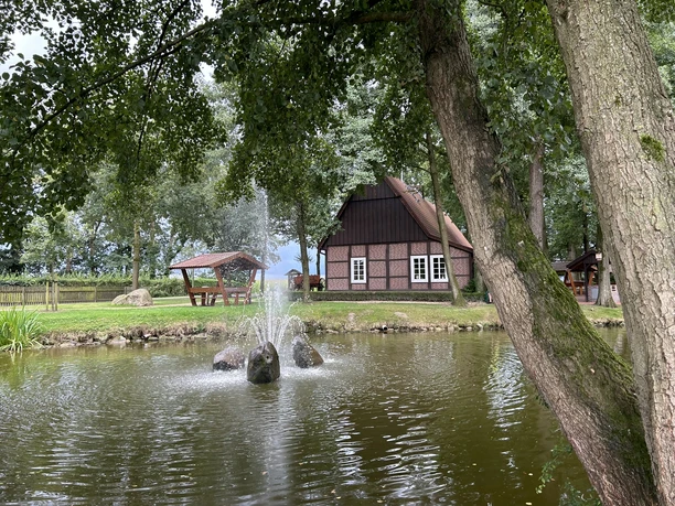 Fachwerkhaus am Teich mit Springbrunnen, umgeben von Bäumen und Sitzbank unter hölzerner Überdachung.